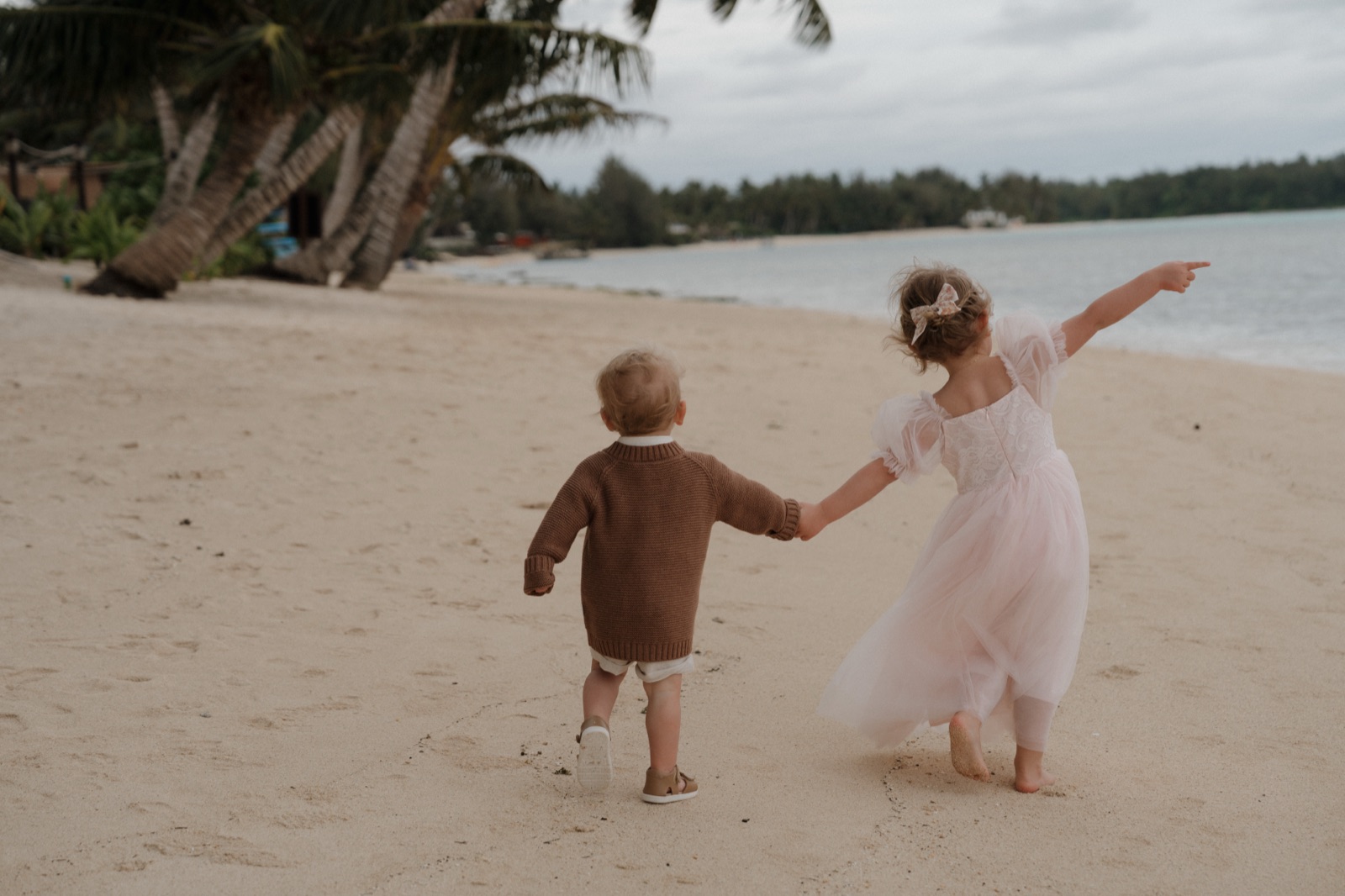 A flower girl and a toddler holding hands on the sand