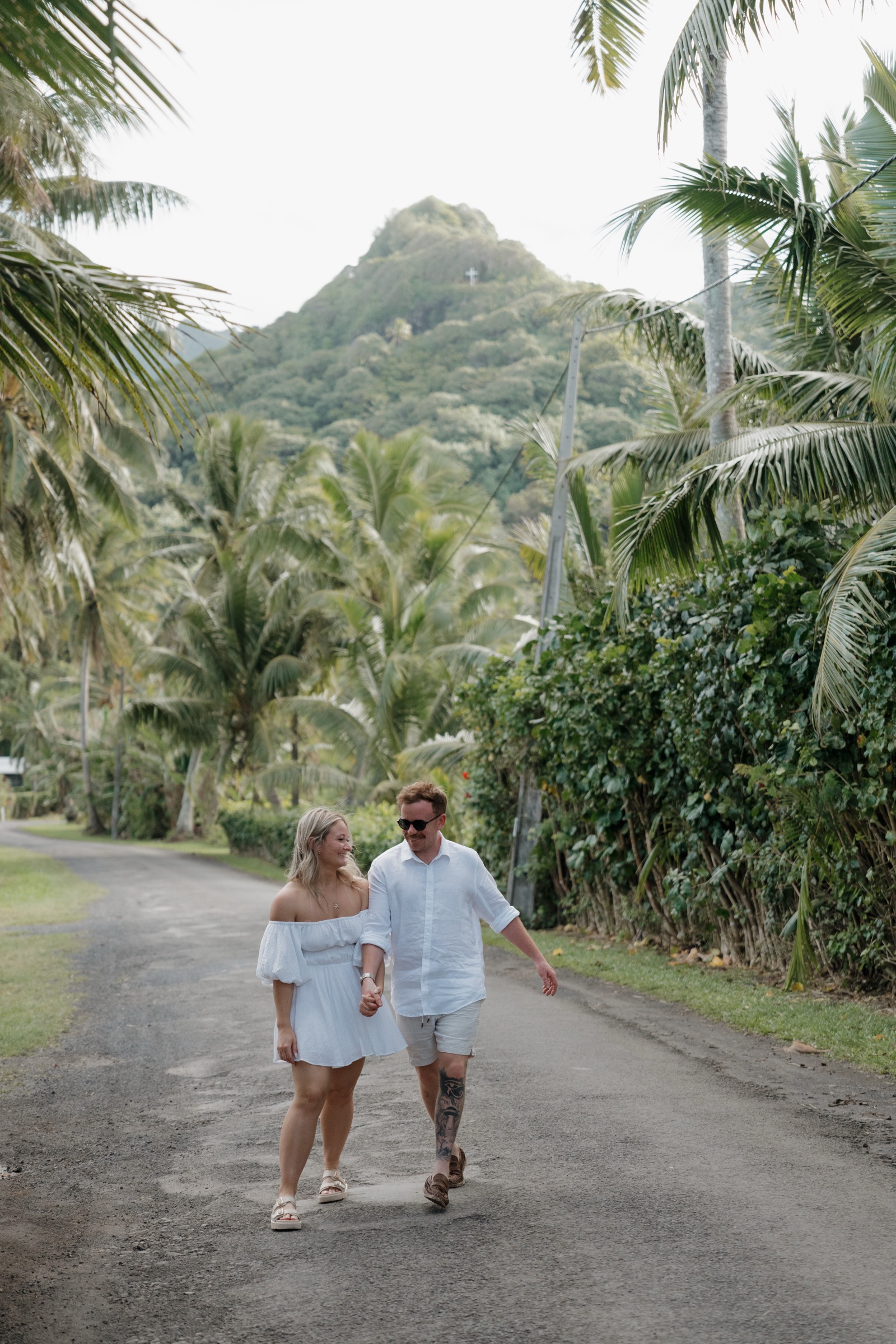 A couple walking hand in hand down a palm-lined island road, a green peak rising behind them