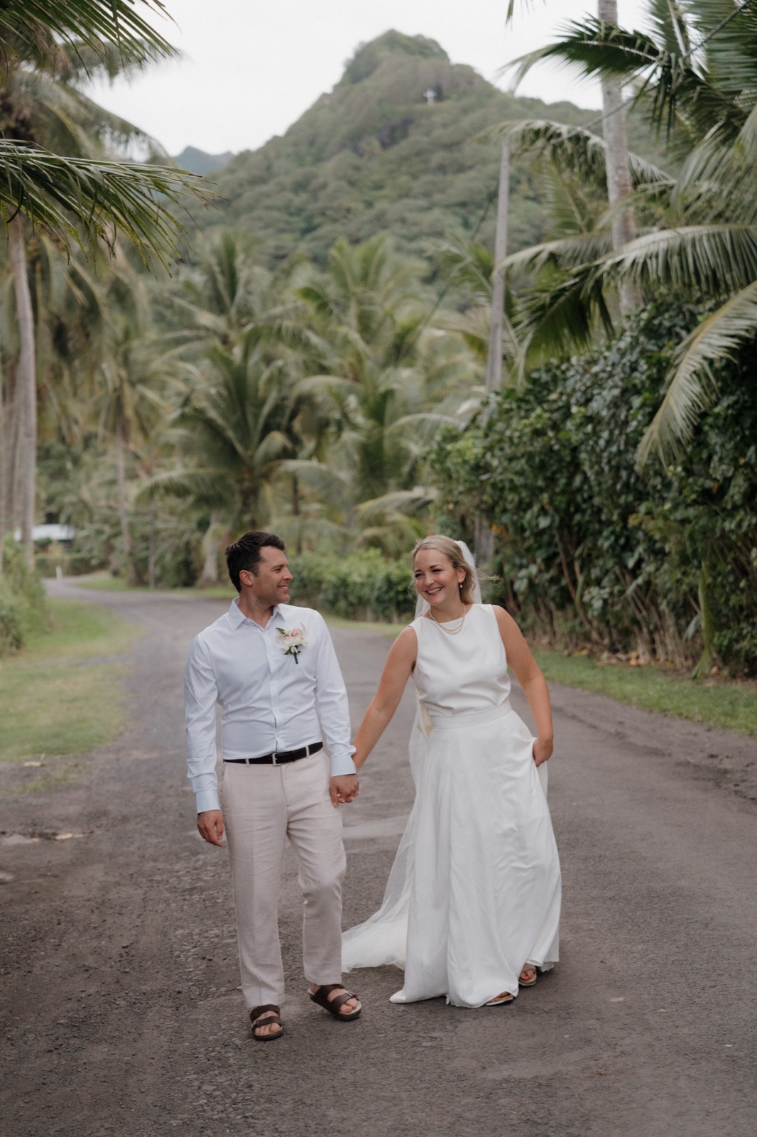 A bride and groom walking hand in hand down a palm-lined road